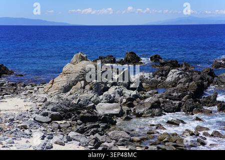 Felsige Küste mit Wellen und fernen Bergen am Strand Santa Maria di Ricadi in Kalabrien unter klarem blauem Himmel in Süditalien. Stockfoto