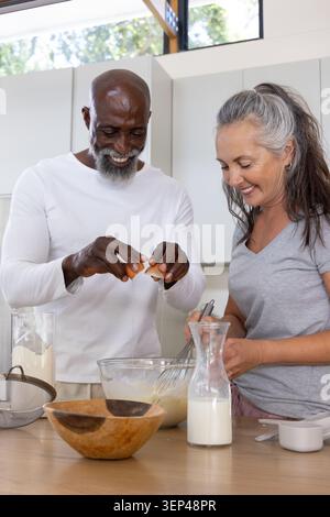 Ein älterer afroamerikanischer Mann, der Eier in eine Glasschale knackt, eine reife Frau, die auf die Insel rauscht Stockfoto