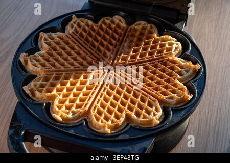 Frisch gebackene Waffeln in Herzform im elektrischen Waffeleisen mit warmem Sonnenlicht, das von einem Seitenfenster über die Oberfläche strahlt Stockfoto