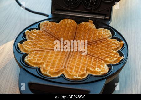 Frisch gebackene Waffeln in Herzform in einer elektrischen Waffelmaschine. Traditionelles, hausgemachtes skandinavisches Dessert, das warm und goldbraun serviert wird Stockfoto