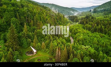 Bergwaldtal mit einem Schutz am Flussufer und Wanderwegen, die von Drohnen gesehen werden, während dünne Nebel durch Nadelbäume driften und sanfte Bergrücken in die Ferne zurückfallen Stockfoto