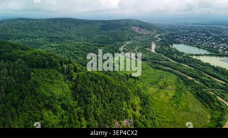 Blick aus der Vogelperspektive auf üppige grüne Hügel und ein breites bewaldetes Tal mit einer gewundenen Straße neben einem ruhigen See und einer kleinen Stadt unter einem weichen bewölkten Himmel Stockfoto