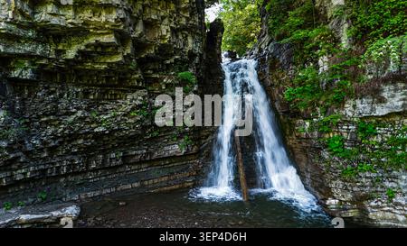 Ein kleiner Wasserfall teilt sich über Grundgestein und fällt in ein flaches Becken innerhalb eines zerklüfteten Canyons. Grüne Pflanzen kleben an Vorsprüngen, während der lebhafte Fluss sprudelt Stockfoto