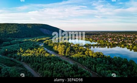 Grüne Wälder umgeben einen reflektierenden See und einen schlängelnden Fluss neben einer kleinen Stadt. Von einer Drohne aus gesehen, zeigt die Szene Sommerlaube Wasserwege und sanfte Bäche Stockfoto