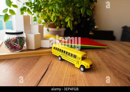 Spielzeug Schulbus auf hölzernem Schreibtisch mit farbenfrohen Schreibwaren Büroklammern Notizbücher und Zimmerpflanzen schaffen ein helles Back-to-School-Thema für den Unterricht Stockfoto