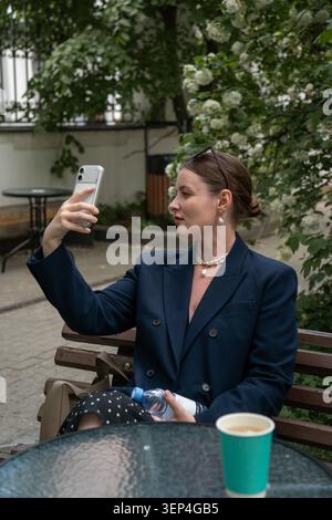 Die junge Frau sitzt auf der Terrasse des Cafés im Freien und hält ein Smartphone für Selfie, während sie sich mit einer Wasserflasche im grünen Innenhof, umgeben von blühender Blüte, ausruht Stockfoto