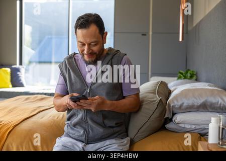 Asiatischer Mann, der auf der Bettkante in einem modernen Schlafzimmer sitzt und das Smartphone in der Nähe einer Senfdecke hält Stockfoto