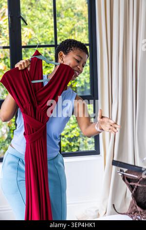Erwachsene afrikanische Frau trägt hellblaues Oberteil und hält zu Hause kastanienbraunes Kleid am offenen Koffer Stockfoto