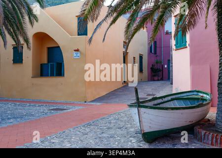 Eine malerische Szene auf einem pulsierenden mediterranen Dorfplatz mit einem alten Fischerboot, farbenfrohen Gebäuden mit grünen Fensterläden und umrahmter Palme Stockfoto