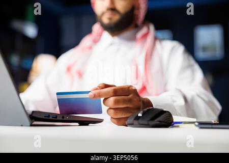 Selektiver Fokus auf einen Mann aus dem Nahen Osten, der eine Debitkarte für nahtloses Online-Shopping ergreift. Nahaufnahme eines arabischen Mannes in traditioneller Kleidung, der mit seinem Computer bezahlt. Stockfoto