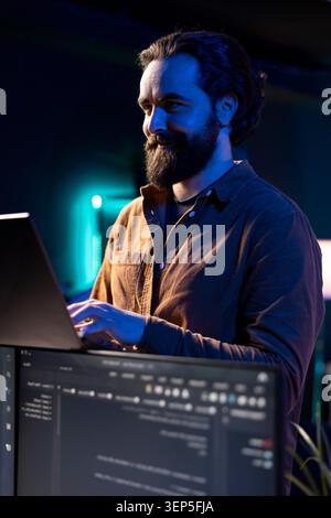 Fröhlicher Freelancer, der Software-Debugging mit einem Laptop, Fehler in Codezeilen auf PC-Monitoren prüft, fröhlicher IT-Profi in der Wohnung läuft und Codierungsprobleme mit Notebook behebt. Stockfoto