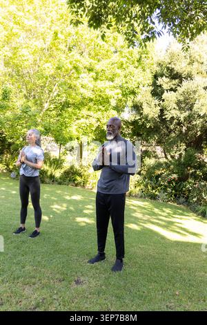 Paare, die Yoga üben, stehen auf grasbewachsenem Rasen unter Bäumen und tragen sportliche Kleidung Stockfoto