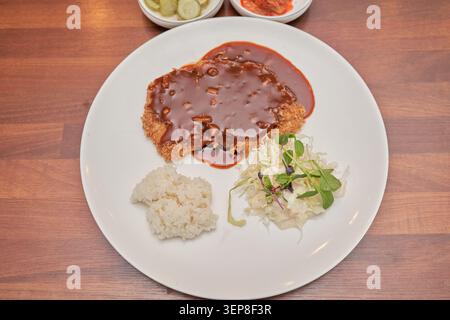 Blick von oben nach unten auf frittiertes Schweinekotelett, bedeckt mit dunkelbrauner Sauce auf einem weißen Teller mit gedämpftem Reis und Kohlsalat über einem Holztisch Stockfoto