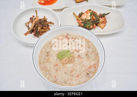 Asiatischer Muschelbrei mit grünem Pulver und einem Löffel Reis und Meeresfrüchten mit verschwommenem scharfem Kimchi auf einem weißen Tisch, der gesunde regionale Di zeigt Stockfoto