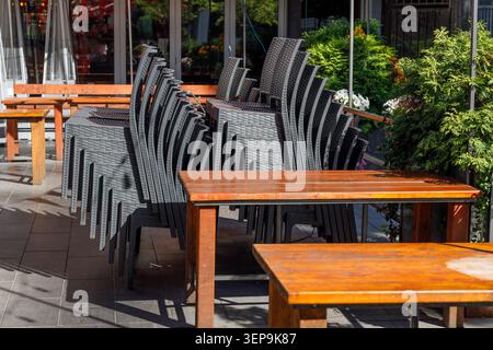 Lackierte Holztische und -Bänke sitzen neben Reihen von gestapelten schwarzen Korbstühlen auf einer ruhigen Terrasse mit hängendem Blumenkorb und üppigem gre Stockfoto