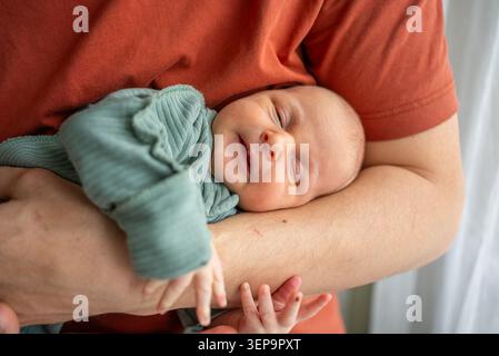 Vater, der seinen neugeborenen Sohn in einer zärtlichen Umarmung umarmt, das Baby schläft friedlich vor der Brust des Väters, fängt Wärme, Vertrauen und die intime Bindung der frühen Elternschaft ein Stockfoto