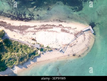 Diese atemberaubende Luftaufnahme fängt eine ruhige Strandszene mit weichem weißem Sand, üppigem Grün und einem hölzernen Pier in klarem Türkis ein Stockfoto