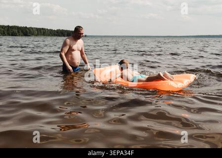 Vater und Sohn genießen einen entspannten Sommertag, schwimmen auf einer aufblasbaren Matratze in einem ruhigen Fluss. Der Mann hält das Kind sicher fest Stockfoto