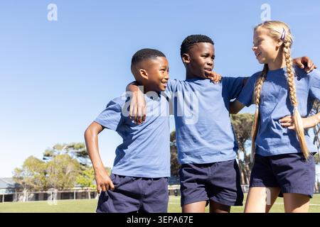 Verschiedene Schulkinder, die ihre Arme miteinander verbinden und sich auf dem Schulfeld unterhalten, tragen blaue T-Shirts und marineblaue Shorts Stockfoto