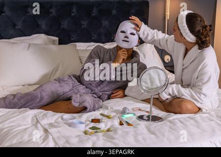 Ein Paar, das sich im Schlafzimmer auf dem Bett liegend, Bademäntel trägt und eine LED-Gesichtsmaske mit Jaderollen reguliert Stockfoto