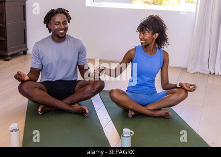 Afroamerikanisches Paar, das Hände auf Matten hält und Wasserflaschen in Sportbekleidung zu Hause hat Stockfoto