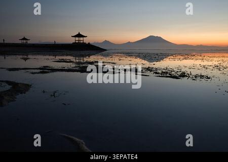 Atemberaubende Reflexion des Vulkans Mount Agung und eines traditionellen balinesischen Pavillons auf der glasklaren, ruhigen Meeresoberfläche während eines friedlichen Sonnenaufgangs in Sanur Bali Stockfoto