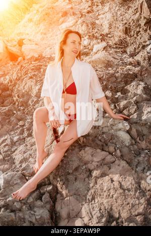 Frau Strand Felsenküste sitzt auf Klippen und trägt roten Bikini und weißes Hemd bei Sonnenuntergang für Sommerurlaub Vibes Stockfoto