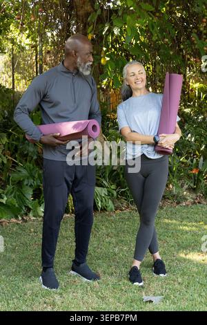 Verschiedene Paare stehen auf dem grasbewachsenen Hof mit gerollten lila Yogamatten, während sie eine Fitnessuhr tragen Stockfoto