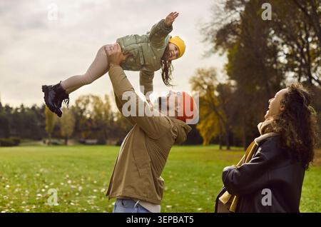 Vater hebt die Tochter im Herbstpark, während Mutter lächelt, fröhliches Familienspiel Stockfoto