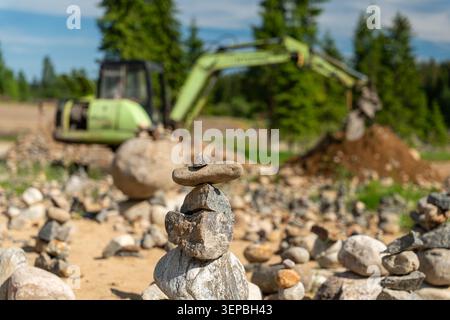 Ein kleiner Stapel ausgeglichener Steine steht im scharfen Fokus auf sandigem Boden, mit verschwommenem Bagger, Felsen und immergrünen Bäumen im Hintergrund im Freien Stockfoto