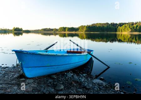 Ein kleines blaues Ruderboot liegt an einem felsigen Seeufer neben einem stillen See, mit sanftem Sonnenaufgangslicht, Baumreflexen, offenem Himmel und einer friedlichen natürlichen Umgebung Stockfoto