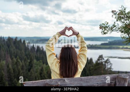 Rückansicht einer Frau in gelbem Pullover, die mit ihren Händen Herzform bildet, während sie mit einem malerischen Blick über einen ruhigen See und einen immergrünen Wald unter sich steht Stockfoto
