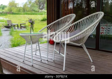 Zwei weiße gewebte Lounge-Stühle und ein kleiner runder Tisch mit einer Glasvase mit Blumen auf einer Holzterrasse neben einem Haus, mit grünem Garten und im Freien Stockfoto