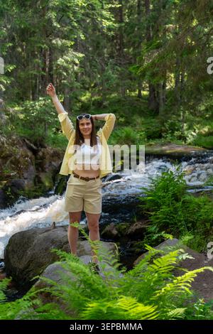 Junge Frau posiert auf großen Felsen in der Nähe des fließenden Flusses im üppigen grünen Wald in lässiger Sommerkleidung und lächelt im hellen Tageslicht mit Farnen Stockfoto