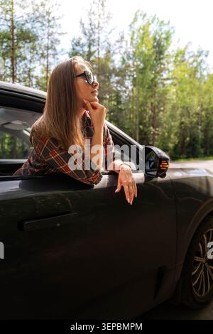 Eine junge Frau mit Sonnenbrille beugt sich an einem sonnigen Tag aus einem schwarzen Autofenster, legt ihr Kinn auf ihre Hand, mit Bäumen und offener Straße hinter ihr Stockfoto