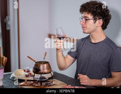 Junger Mann, der ein Glas Rotwein hält und das Aroma genießt, bevor er ein traditionelles Fleischfondue Bourguignonne genießt, das in kochendem Öl mit verschiedenen Zutaten zubereitet wird Stockfoto