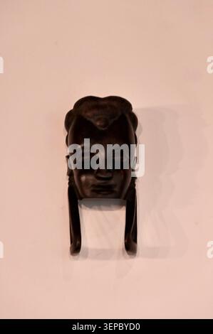 Eine auffällige vertikale Nahaufnahme einer dunkelbraunen, geschnitzten afrikanischen Holzmaske oder eines ethnischen Figurenkopfes vor einem sauberen beigefarbenen Hintergrund. Die dra Stockfoto