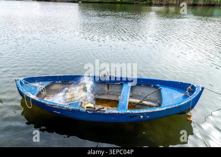 Ein kleines blaues Fischerboot mit sichtbarer Abnutzung schwimmt auf ruhigem Wasser, mit Fischernetz, Seil und einem Holzpaddel im Rumpf. Stockfoto