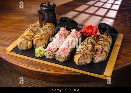 Sushi-Brötchen werden auf eine Holzplatte gelegt. Es gibt verschiedene Arten von Brötchen mit Meeresfrüchten und Belägen. Dippsoße und Beilagen sind neben dem Sushi. Die Umgebung verfügt über Tageslicht. Stockfoto