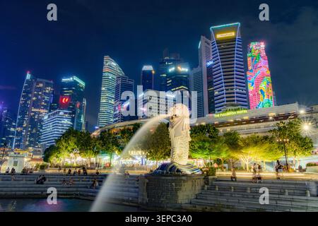 Nächtlicher Blick auf die Merlion-Statue, die Wasser spuckt, mit der beleuchteten Skyline des zentralen Geschäftsviertels von Singapur im Hintergrund. Stockfoto