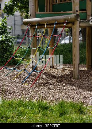 Eine farbenfrohe Seilleiter hängt an einem hölzernen Spielplatzrahmen in einem Park im Freien mit Mulch-Bodenabdeckung, grünem Gras, Bäumen und Apartment-Gebäude Stockfoto