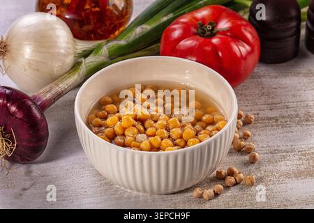 Weiße Schüssel gefüllt mit gekochten Kichererbsen in Brühe, umgeben von roten Zwiebeln, weißen Zwiebeln, Tomaten, grünen Zwiebeln und Gewürzen auf strukturierter Oberfläche Stockfoto