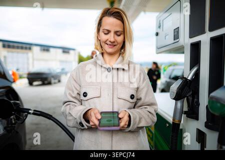 Frau, die an der Tankstelle mit Smartphone für Treibstoff bezahlt Stockfoto