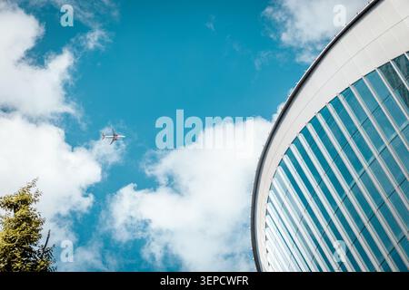 Abstrakte Ansicht des flachen Winkels der geschwungenen Glas- und Stahl-Wolkenkratzerfassade mit einem kommerziellen Flugzeug, das vor blauem Himmel und weißen Wolken vorbeifliegt, Taipeh Stockfoto