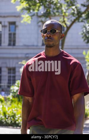 Afroamerikaner, der mit einer dunklen ovalen Sonnenbrille und einem kastanienbraunen Hemd am Steingebäude steht Stockfoto
