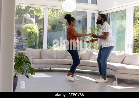 Afroamerikanisches Paar tanzt und hält Hände in einem modernen Wohnzimmer an raumhohen Fenstern Stockfoto