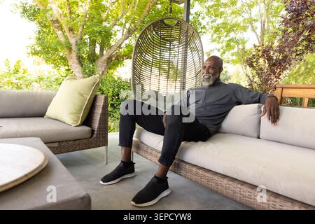 Ranghoher afroamerikanischer Mann, der auf Rattan-Veranda-Sofa in dunklem Pullover neben hängendem Eierstuhl sitzt Stockfoto