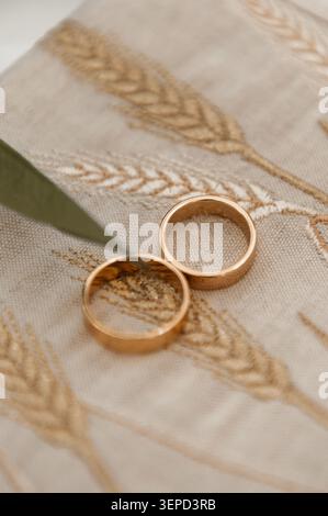 Eine detaillierte Nahaufnahme eines Paares klassischer goldener Eheringe auf einem hellen Leinenstoff mit zarter goldener Weizenstickerei. Stockfoto