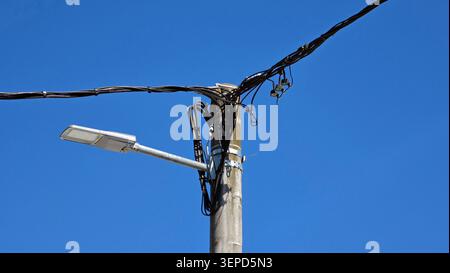 Eine moderne LED-Straßenleuchte ist an einem Betonmast montiert, der mit Bündeln dicker schwarzer Stromkabel und Überlandleitungen verwickelt ist Stockfoto