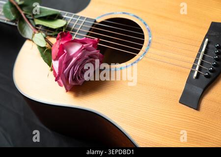 Eine rosa und rote Rose, die auf einer Akustikgitarre ruht. Ein romantisches Stillleben, das Musik, Liebe und künstlerischen Ausdruck mit satten Texturen, warmen Holztönen und dramatischen Kontrasten kombiniert. Stockfoto